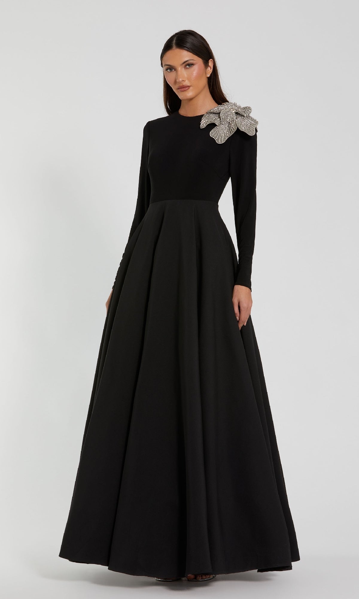 A woman wears the Mac Duggal 11832, a long sleeve black formal dress with an A-line silhouette and a large silver embellishment on one shoulder, standing against a plain light background.